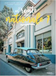 Couverture du livre En route… sur la nationale 7 avec une voiture ancienne noire stationnée devant un bâtiment rétro aux teintes vert pastel et blanc, ambiance estivale et vintage, titre en typographie manuscrite jaune. Achat sur Divertistore.com.
