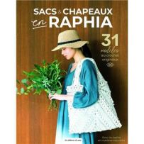 Couverture du livre Sacs & Chapeaux en Raphia, montrant une femme vêtue d’une robe ample bleue et portant un grand sac crocheté en raphia blanc ainsi qu’un chapeau assorti. Elle tient un bouquet de verdure, sur fond en bois naturel. Le titre indique « Sac
