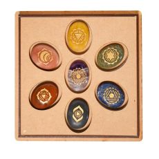 Photographie du "Coffret Galets 7 Chakras" ouvert. On y voit sept galets ovales en pierres naturelles de différentes couleurs (améthyste, lapis-lazuli, jaspe, etc.), bien rangés dans des emplacements ronds sur un fond cartonné beige. Chaque pierre est orn